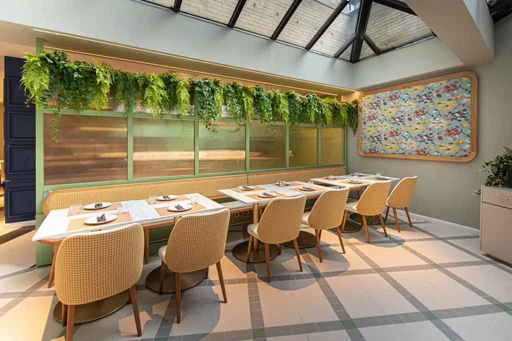 Heller Restaurantbereich mit Glasdach, weißen Tischen, gelb-karierten Stühlen, grüner Hängepflanzen-Dekoration und einem bunten Wandbild